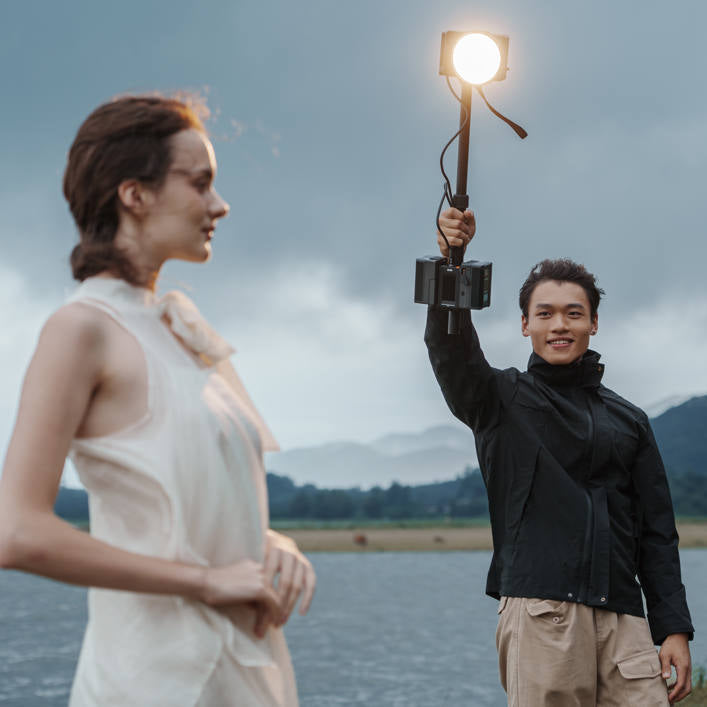 Person holding a lamp near another person by a body of water with mountains in the background