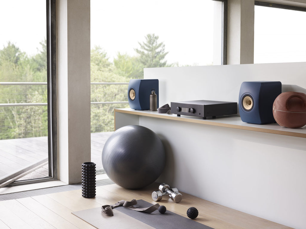 KEF LS50 Meta passive bookshelf speakers in Royal Blue, shown on a shelf in an exercise room setting.