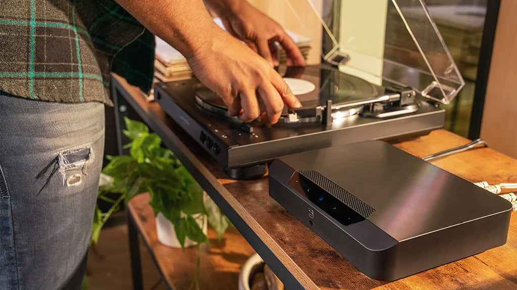 Bluesound Powernode Edge music streamer in black, shown connected to audio equipment in a modern living room setup.
