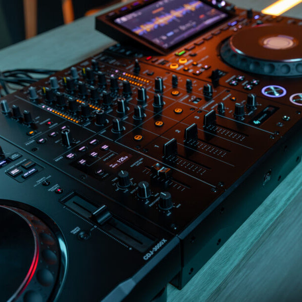 AlphaTheta DJM-V5 DJ mixer with professional controls, display and sleek black design, shown in a lifestyle setting.