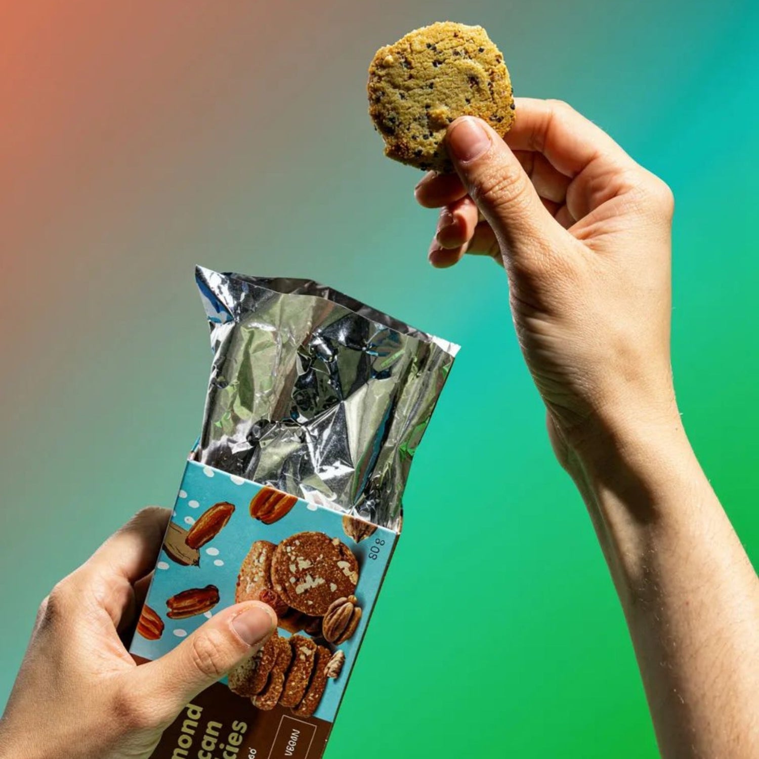 Hand holding a cookie above a packet of almond pecan cookies against a gradient background